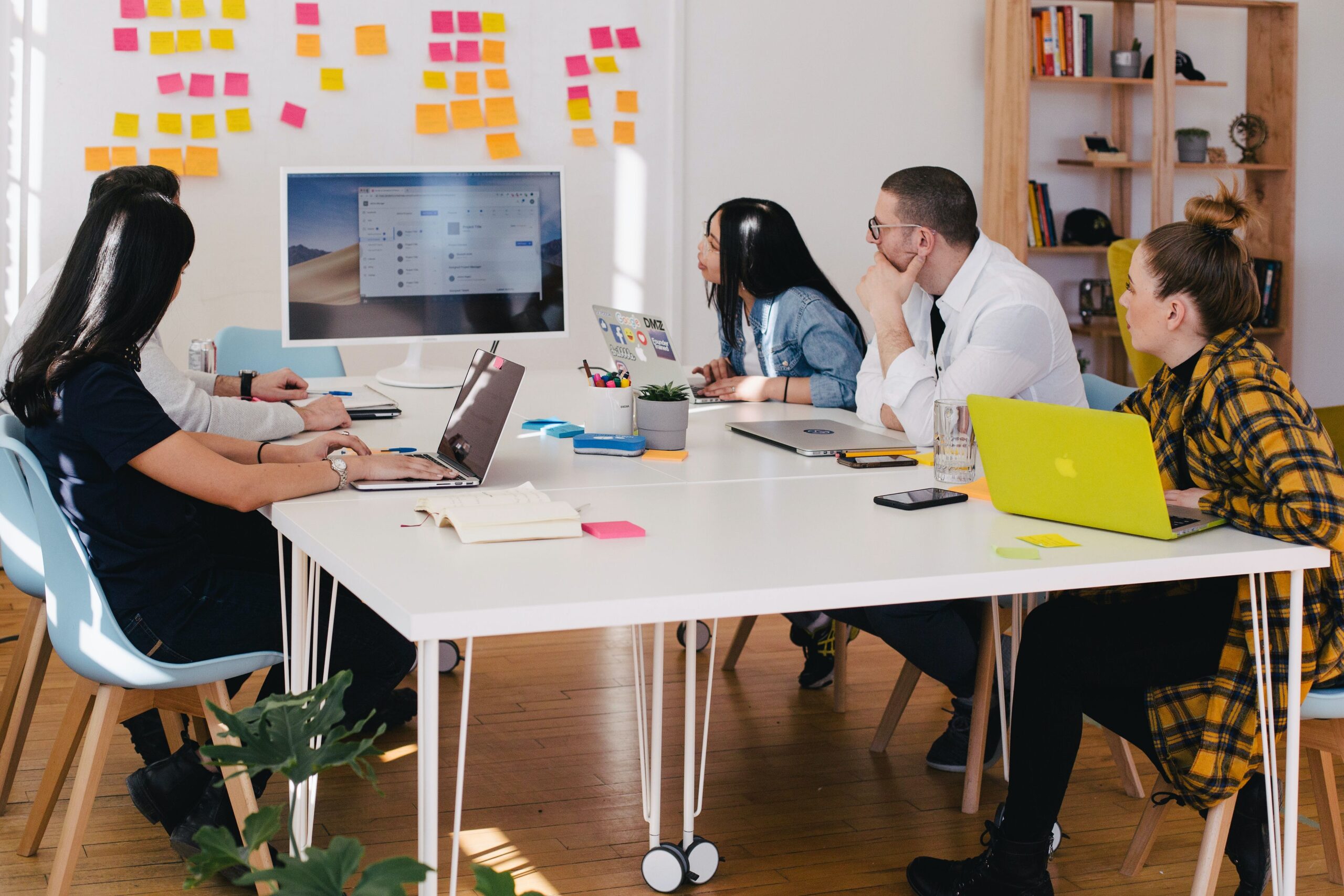 Foto einer Team Besprechung, alle sitzen an einem weißen Tisch mit Computern vor sich und schauen auf einem großen Monitor am Ende des Tisches.