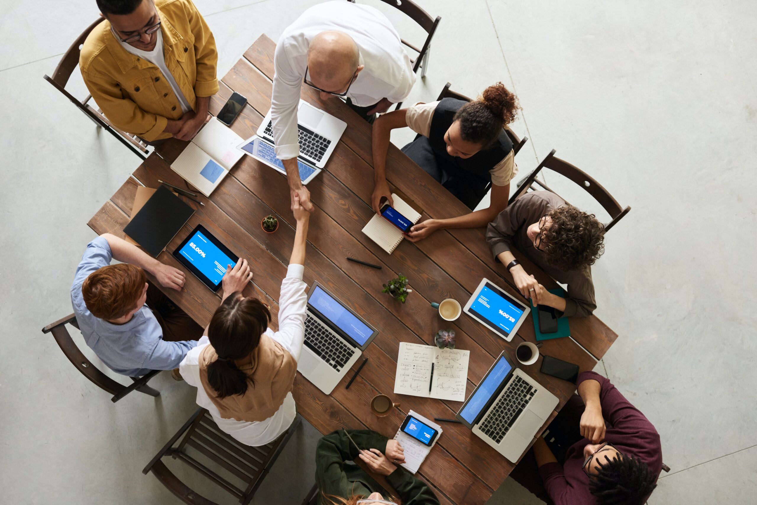 Foto eines Team Meetings mit 8 Personen aus der Vogelperspektive, alle sitzen an einem kleinen Holztisch mit Dokumenten, Computern und Tablets, wobei 3 Teammitglieder aufgestanden sind und zwei sich die Hände reichen.